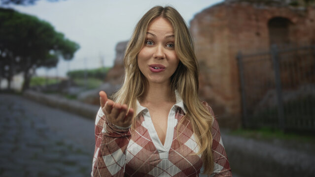 Woman with uncovered hand waves and speaks to camera at city ruins; greeting connection friendliness warmth.