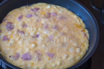 Close-up shot of an egg mixture with ham and onion pieces cooking in a black non-stick frying pan. The omelet is still partially liquid with visible bubbles