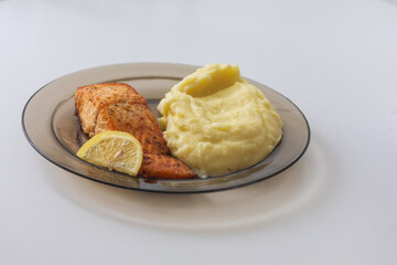A portion of baked salmon fillet with a lemon wedge and a dollop of mashed potatoes, served on a round, tinted glass plate on a white table