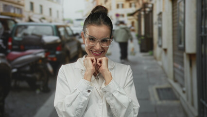 Fototapeta premium Hispanic woman scientist wearing lab coat smiling broadly with fists under chin on city street; optimism.