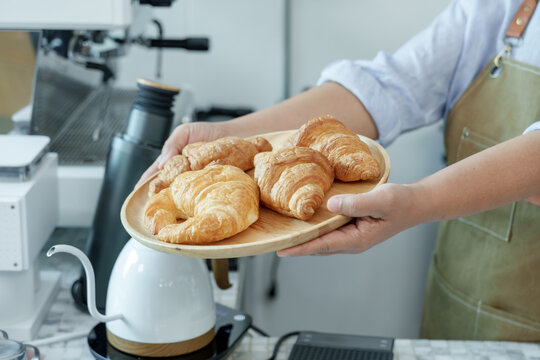Close-up of Asian elderly barista hands holding wooden plate of croissants near coffee equipment, reflecting craftsmanship, passion for baking, hospitality, charm of cozy cafe.