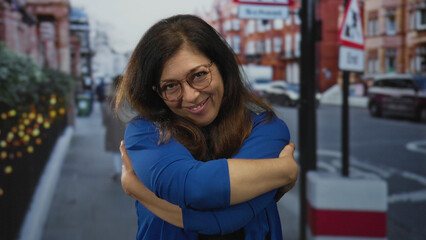 Woman wearing glasses and a blue shirt smiling while hugging herself on a busy urban street in daytime; self acceptance.