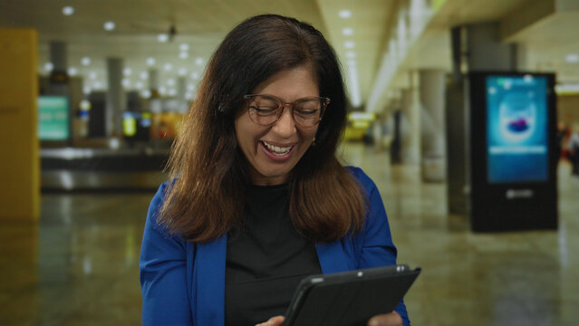 Middle aged hispanic woman holds tablet over face in busy airport terminal, smiling broadly; excitement.