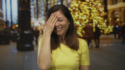 Fototapeta premium Hispanic middle aged woman places hand to forehead with a slight smile on street illuminated by decorative lights; embarrassment.