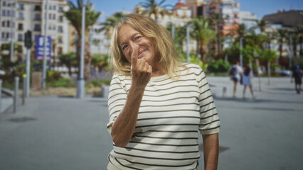 Senior woman in a striped shirt shows middle finger on a sunny street promenade with palm trees and passing pedestrians; defiance.
