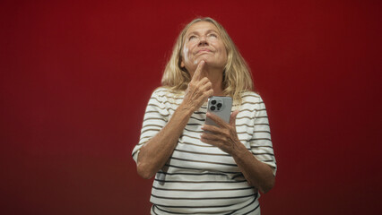 Senior woman holding smartphone and pointing finger to chin while smiling in a studio; quiet...