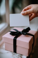 Female hand holding blank card over pink gift box with black ribbon, clean aesthetic, soft focus