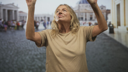 Senior blonde woman raising both arms with bare forearms visible in a busy piazza before a basilica, looking upward  quiet contemplation. © Krakenimages.com