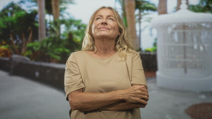 Senior woman with arms crossed and chin lifted, sometimes pointing finger, standing by a white fountain on a palm lined street, casual beige shirt and relaxed posture; calm reflection.