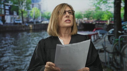 Woman judge in black robe with glasses reading a legal brief by canal street outdoors in city scene...