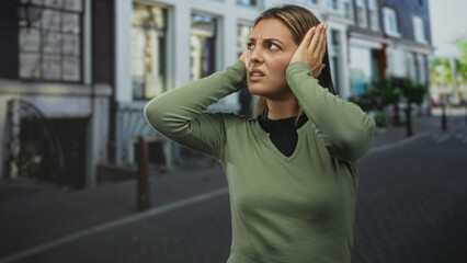 Young woman covering ears with hands on a city street, grimacing and squinting against loud noise; sensory overload pain.