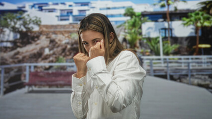 Young blonde woman throwing a punch with fists raised in front of a building on a boardwalk with palms and railing; determination resilience.