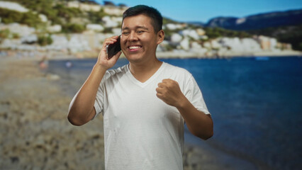 Hispanic man talking on phone at beach, smiling and holding smartphone with raised fist gesture; joy connection.