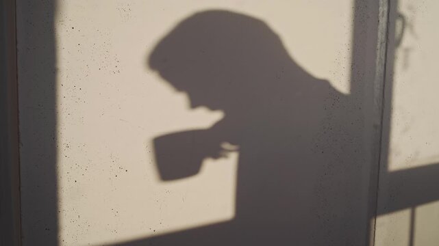 The shadow of a person with a disability is seen on a balcony as they quietly drink coffee at dawn.
