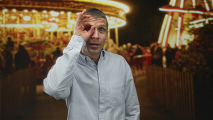 Fototapeta premium Man making gesture in front of eye at night fair with carousel lights creating vibrant outdoor atmosphere.