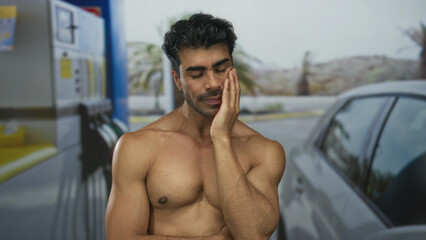 Shirtless man holds cheek at a petrol station outdoors in sunlight with a parked car nearby and palm trees beyond  fatigue. © Krakenimages.com