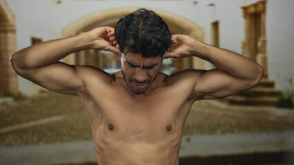 Obraz premium Shirtless young hispanic man covering ears on deserted street lined with old stone buildings; discomfort.