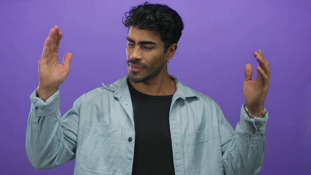 Young hispanic man in a light blue denim shirt holding hands apart measuring width in a purple studio; size evaluation.