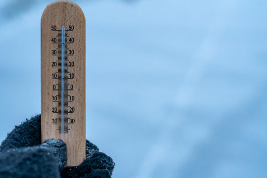 Light frost shown on a wooden thermometer held in a glove against a snowy winter landscape backdrop