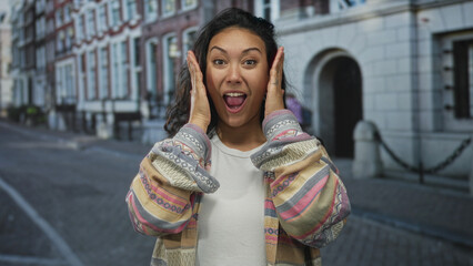 Fototapeta premium Woman holding hands to face and smiling on cobblestone city street with row houses and stone building facades visible behind her; playful surprise.