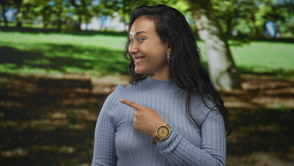 Woman smiling and pointing finger at a tree in a bright forest park with green grass and a bench,...