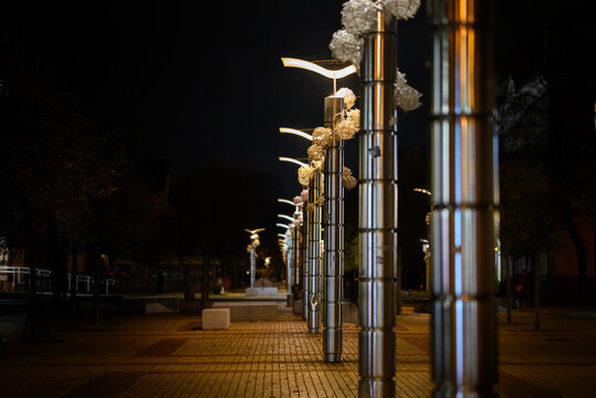 Tall metal poles with modern streetlights line a paved walkway at night. Each pole is adorned with decorative metallic spheres, enhancing the futuristic appearance.