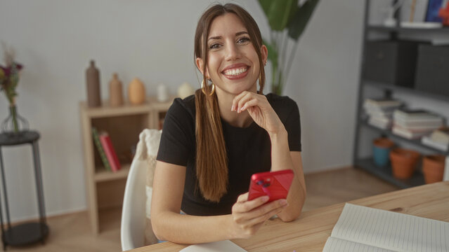 Young hispanic woman holding red smartphone and resting hand on chin in building interior; contemplation reflection introspection mindfulness. - Powered by Adobe