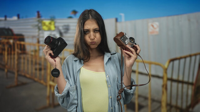 Woman holds camera in each hand at construction site, smiling and showing vintage and mirrorless cameras; joy creativity exploration. - Powered by Adobe