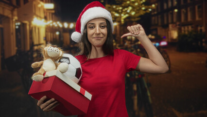 Woman wearing santa hat holding red gift box and soccer ball, thumb pointing to self on night...
