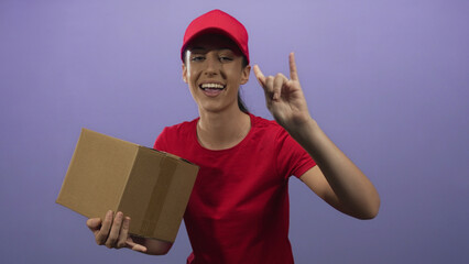 Woman holding cardboard box and making rock horn hand gesture in studio purple; friendly delivery...