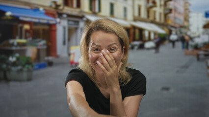 Woman with blonde hair in black shirt smiles broadly and points finger at camera on busy street; joy playfulness.