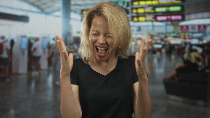 Blonde woman with raised hands shouting in a busy airport terminal near departure board under glass...