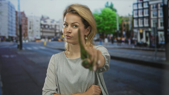 Blonde woman points her finger forward at busy city street intersection wearing gray blouse; bold confidence.