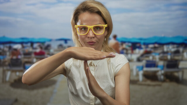 Young blonde woman with yellow glasses makes timeout sign on sandy beach amid blurred umbrellas; frustration.