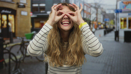 Woman raises hands gesturing on a street with binoculars, appearing blonde, smiling, playful, urban, happy.