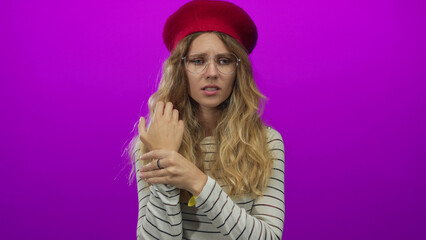 Naklejka premium Image captures woman inspecting her wrist and examining in a studio where blonde young style meets beret, sweater, glasses on a purple background.