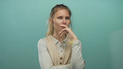 Woman touches finger to lips while thinking in studio with blonde young pensive portrait staged against teal background.