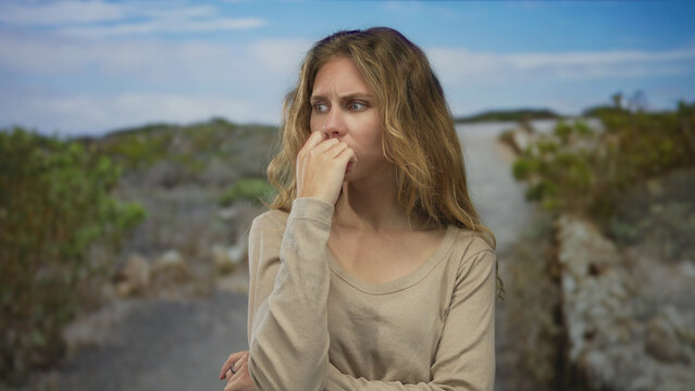 Woman with finger biting gesture outdoors shows a blonde young anxious posture as she presses nail against mouth on a rocky path trail.