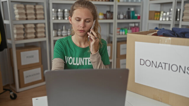 Woman holds phone while calling from a warehouse office, typing on laptop as volunteer organizes donations on a shelf beside a box and checks inventory.
