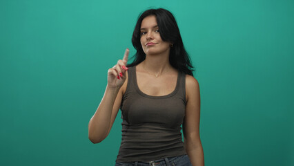 Young hispanic woman pointing index finger upward against a bright teal studio wall with direct gaze; confidence.