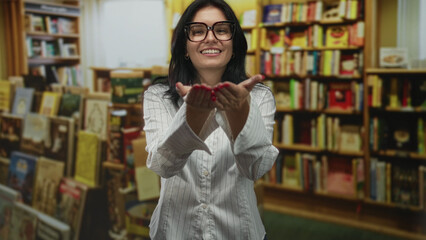 Woman wearing glasses and a white striped shirt blowing a kiss with her hands amid bookstore shelves; connection intimacy closeness affection.