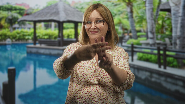Woman points index finger and taps other hand by a pool building gazebo, wearing floral blouse and glasses; friendly instruction.