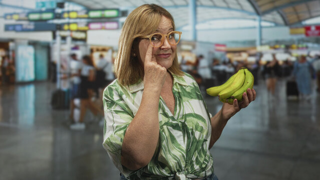 Middle age woman touching eyeglasses with fingertip while holding banana in airport terminal; playful curiosity.