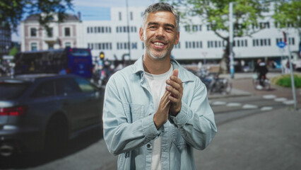 Man clapping hands on a busy street beside parked car and cyclists, middle age grey hair wearing light jacket with closed eyes in mid gesture; contentment.
