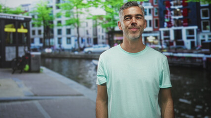 Man in green tshirt raises hands palms forward and sticks out tongue on a canal street; playful...