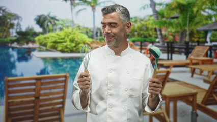 Fototapeta premium Man chef in white jacket, middle age with grey hair, holds whisk and ladle by resort poolside, presenting cooking tools to camera; relaxed hospitality.