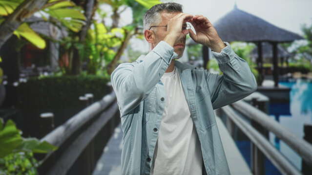 Man forms hands into binoculars and looks along a resort pool walkway across a hotel building bridge; curiosity discovery exploration.
