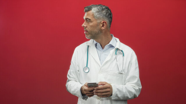 Man doctor in white coat with stethoscope holding smartphone with both hands and looking down at screen, studio; thoughtful. - Powered by Adobe