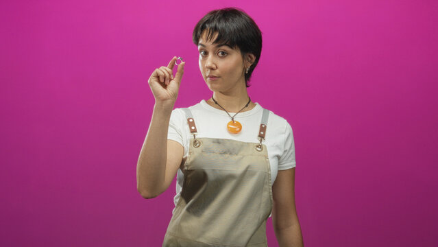Woman jeweler holding a ring between fingers, wearing apron and white tee, short hair, examining the ring while standing in a studio; focused craft.