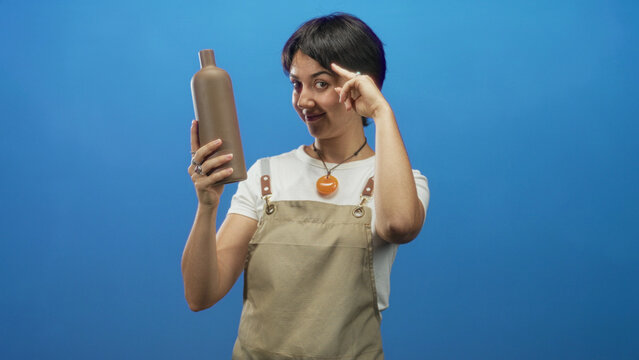 Woman in apron holding a brown pottery bottle with her left hand and pointing two fingers toward camera while smiling in studio; playful charm.